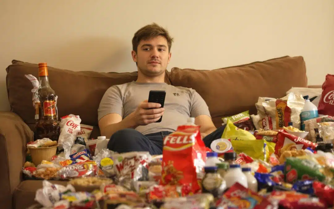 Man on couch surrounded by snacks holding a remote